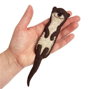 May include: A handmade felt otter in shades of brown and cream. The otter has a smiling face, stitched details, and a long, curved tail. The craft is held in a hand against a white background.