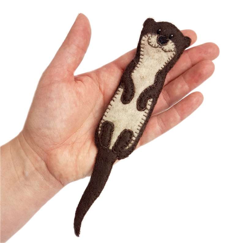 May include: A handmade felt otter in shades of brown and cream. The otter has a smiling face, stitched details, and a long, curved tail. The craft is held in a hand against a white background.