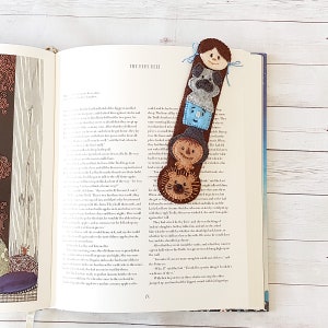 May include: A felt bookmark featuring a stack of five characters, including a girl, a dog, a boy, a scarecrow, and a lion. The bookmark is brown with colorful accents and is perfect for marking your place in a book.