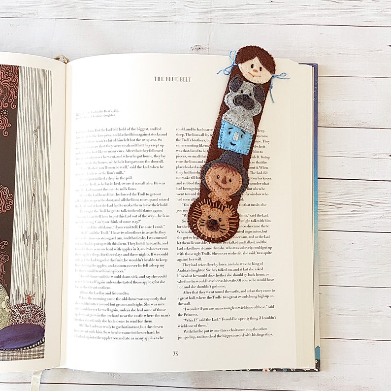 May include: A felt bookmark featuring a stack of five characters, including a girl, a dog, a boy, a scarecrow, and a lion. The bookmark is brown with colorful accents and is perfect for marking your place in a book.