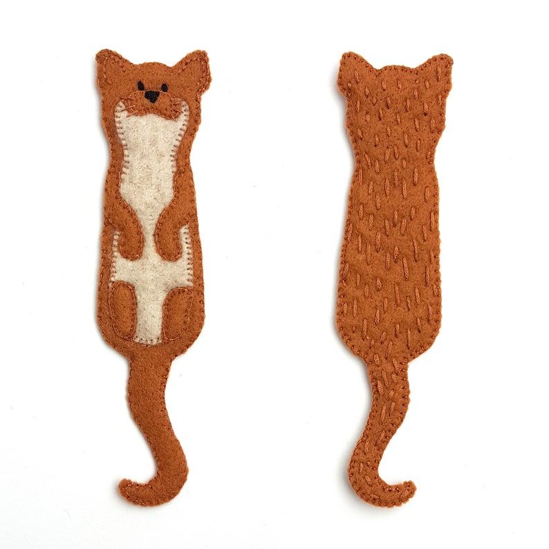 May include: Two felt otter-shaped bookmarks. One shows the front of the otter with a light beige belly and stitched details. The other shows the back of the otter with stitched details. Both are brown with a curved tail.