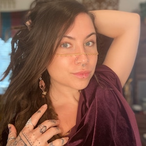 May include: A woman with long brown hair wearing a maroon velvet top and gold jewelry. She has a nose ring and a lip ring. She is wearing a gold chain across her face.