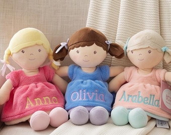 Op de afbeelding: Drie pluche poppen met geborduurde namen op hun jurken. De linkerpop heeft geel haar en een roze jurk met de naam Anna. De middelste pop heeft bruin haar en een blauwe jurk met de naam Olivia. De rechterpop heeft blond haar en een perzikkleurige jurk met de naam Arabella.