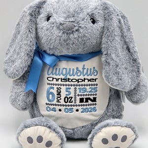 May include: A gray plush rabbit toy with long ears and a blue ribbon tied around its neck. The rabbit features an embroidered white chest panel displaying the name Augustus Christopher, birth weight of 6 pounds 5 ounces, 19.25 inches, and 04-05-2026.
