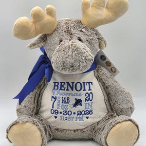 May include: A plush brown moose toy with tan antlers and a blue ribbon around its neck. The moose wears a white chest patch embroidered with the name BENOIT Thomas, 7 pounds 14.5 ounces, 20 inches, 09-30-2026, and 11:57 PM in blue thread.