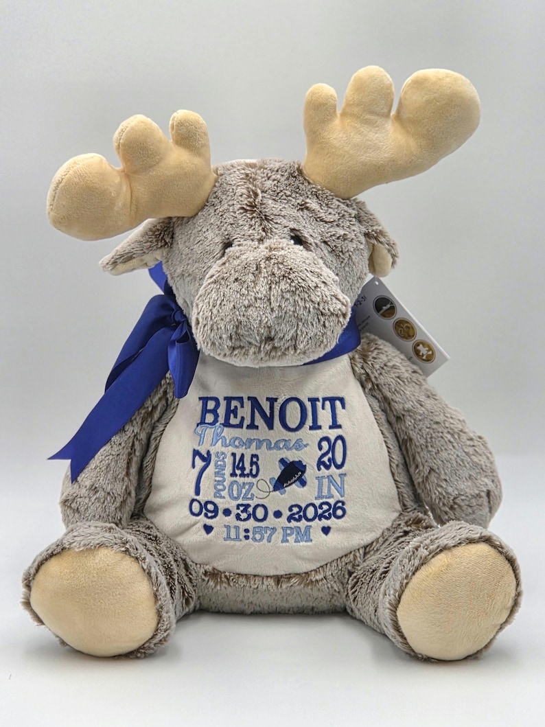 May include: A plush brown moose toy with tan antlers and a blue ribbon around its neck. The moose wears a white chest patch embroidered with the name BENOIT Thomas, 7 pounds 14.5 ounces, 20 inches, 09-30-2026, and 11:57 PM in blue thread.