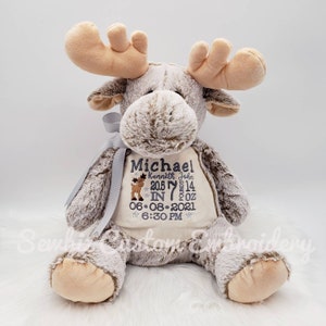 May include: A plush moose toy with light brown fur and tan antlers. The chest features embroidered text: Michael, Kenneth John, 20.5 IN, 7 pounds 14 OZ, 06-08-2021, 6:30 PM, and a small moose graphic. A grey ribbon is tied around the neck.