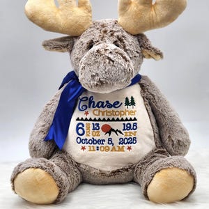 May include: A plush brown moose stuffed animal with a blue ribbon around its neck. The cream-colored belly features embroidered text: Chase Christopher, 6 pounds 13 oz, 19.5 in, October 5, 2025, 11:09 AM, along with mountain and star icons.