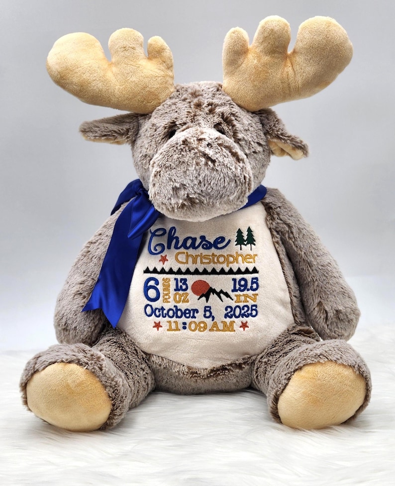 May include: A plush brown moose stuffed animal with a blue ribbon around its neck. The cream-colored belly features embroidered text: Chase Christopher, 6 pounds 13 oz, 19.5 in, October 5, 2025, 11:09 AM, along with mountain and star icons.