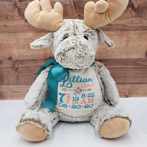 May include: A plush moose toy with soft beige fur and large antlers, wearing a teal satin ribbon around its neck. The chest features embroidered text: Lillian, Davi, 7 pounds, 19 in, 8:22 AM, and 08-20-20, accented with a floral arrow design.