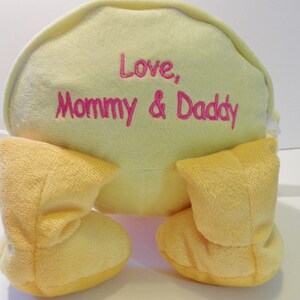 May include: A soft, yellow plush toy with the embroidered text "Love, Mommy & Daddy" in pink. The toy has two yellow, rounded feet. The background is white.