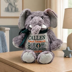May include: A plush gray elephant toy featuring a dark green ribbon around its neck and a personalized embroidered panel on its chest. The embroidery includes the name CALLEN, Edward Szerepanski, birth details 9 pounds 10 oz, 22 1/4 in, and 06-03-2025 04PM.
