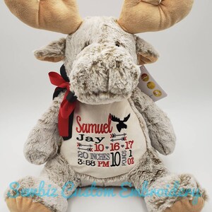 May include: A plush tan moose toy with a red and black ribbon around its neck. The chest features embroidered text: Samuel, Jay, 10-16-17, 20 INCHES, 10 POUNDS 1 OZ, 3:58 PM, along with arrow icons and a small moose silhouette in red and black thread.