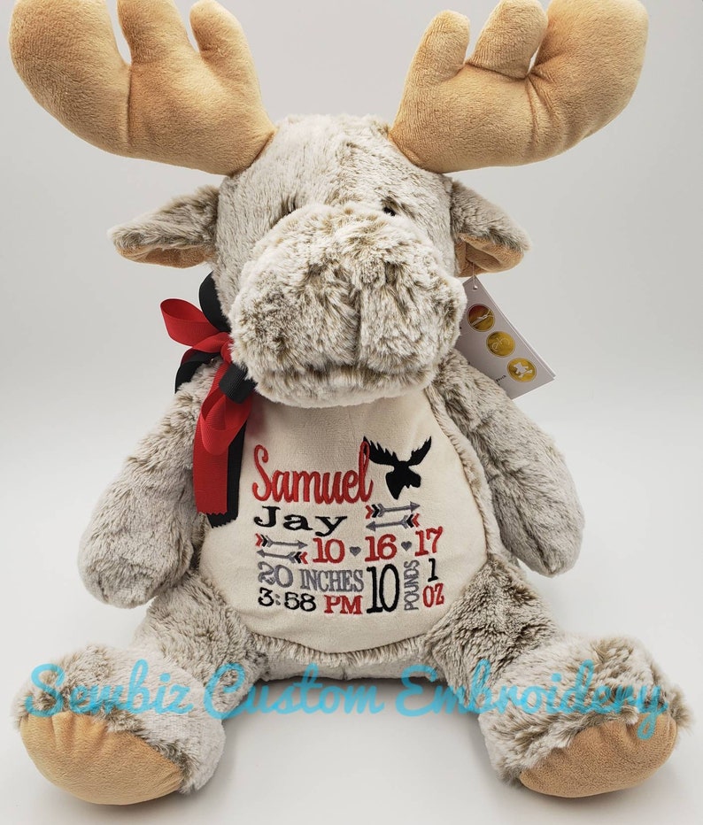 May include: A plush tan moose toy with a red and black ribbon around its neck. The chest features embroidered text: Samuel, Jay, 10-16-17, 20 INCHES, 10 POUNDS 1 OZ, 3:58 PM, along with arrow icons and a small moose silhouette in red and black thread.