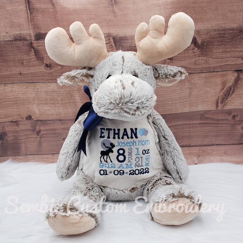 May include: A plush gray moose toy with large beige antlers and a dark blue ribbon around its neck. The chest features embroidered text: ETHAN, Joseph Horn, 8 pounds 1 oz, 21 inches, 9:12 AM, and 01-09-2022, with a moose silhouette and blue hearts.