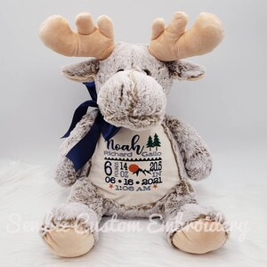 May include: A plush moose toy with light brown fur and tan antlers. A dark blue ribbon is tied around its neck. The belly features embroidered text: Noah, Richard Gallo, 6 pounds 14 oz, 20.5 in, 06-16-2021, 1:06 AM, with mountain and tree icons.