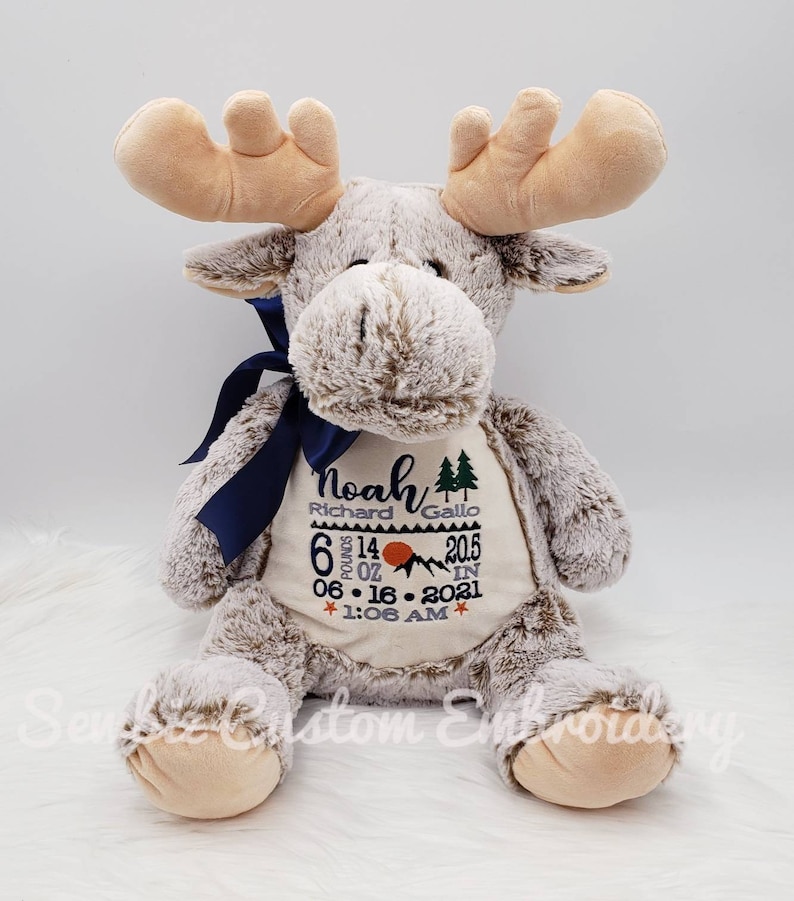 May include: A plush moose toy with light brown fur and tan antlers. A dark blue ribbon is tied around its neck. The belly features embroidered text: Noah, Richard Gallo, 6 pounds 14 oz, 20.5 in, 06-16-2021, 1:06 AM, with mountain and tree icons.