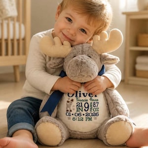 May include: A plush moose toy with soft light brown fur, cream-colored antlers, and a dark blue ribbon around its neck. The white belly features embroidered text: Oliver, Francis Haas, 21 IN, 9 LBS 8.7 OZ, 01-06-2026, 6:12 PM, and a small moose graphic.