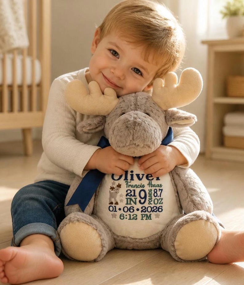 May include: A plush moose toy with soft light brown fur, cream-colored antlers, and a dark blue ribbon around its neck. The white belly features embroidered text: Oliver, Francis Haas, 21 IN, 9 LBS 8.7 OZ, 01-06-2026, 6:12 PM, and a small moose graphic.