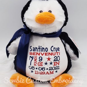May include: A plush penguin toy with black and white fur, an orange beak and feet, and a dark blue satin ribbon around its neck. The white belly features embroidered text: Santino Crue, BENVENUTI, 7 POUNDS 9 OZ, 20 IN, 05.06.2021, and 11:29AM.