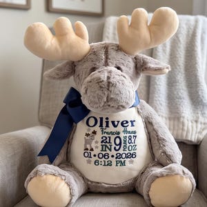 May include: A plush moose toy with soft grey fur and cream-colored antlers. A dark blue ribbon is tied around its neck. The chest features embroidered text: Oliver, Francis Haas, 21 IN, 9 LB 8.7 OZ, 01-06-2026, 6:12 PM, and a small moose icon.