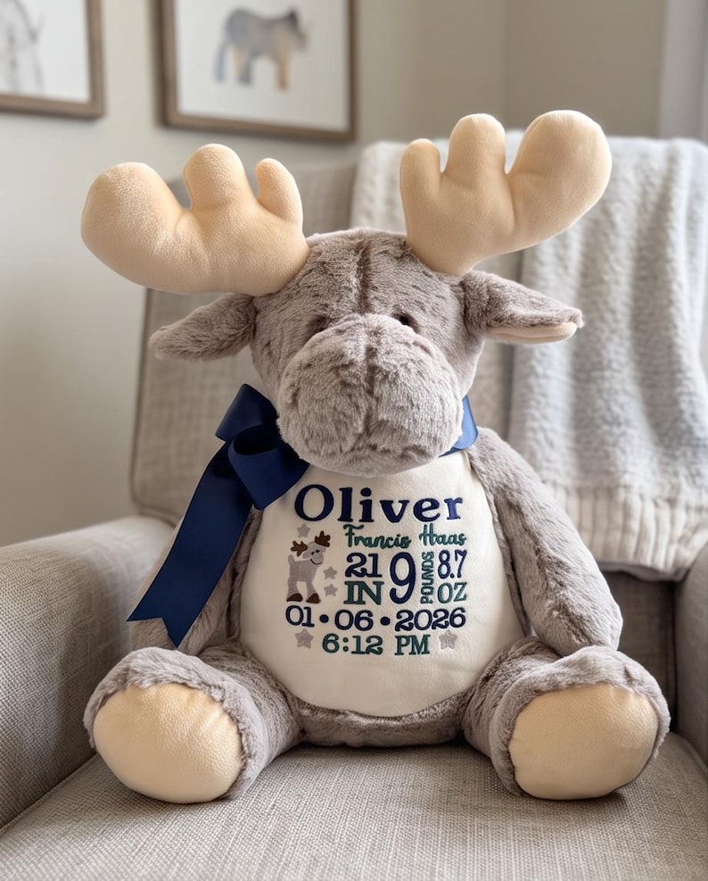May include: A plush moose toy with soft grey fur and cream-colored antlers. A dark blue ribbon is tied around its neck. The chest features embroidered text: Oliver, Francis Haas, 21 IN, 9 LB 8.7 OZ, 01-06-2026, 6:12 PM, and a small moose icon.