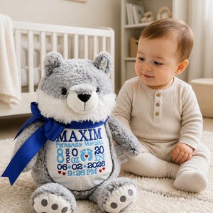 May include: A gray plush wolf toy with a blue ribbon around its neck. The white belly features embroidered text: MAXIM, Fernando Morales, 8 lbs 0 oz, 20 in, 06-02-2026, and 9:26 PM, along with blue baby footprints and small red hearts.