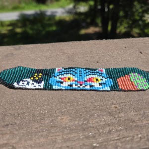 Peut inclure: Un bracelet de perles avec un motif central de chat bleu, flanqué de formes blanches et jaunes d'un côté et d'une section orange et verte de l'autre. Le bracelet est fait de petites perles colorées.