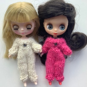 May include: Two Blythe dolls, one with blonde hair and the other with dark hair, are shown. Both dolls wear knitted, long-sleeved jumpsuits. One jumpsuit is cream-colored, and the other is pink. Each jumpsuit has a small, dark button.