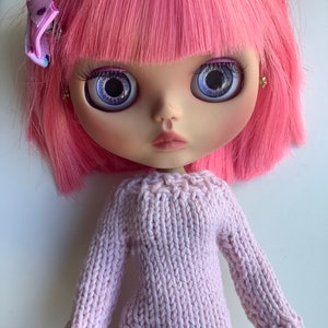 May include: A Blythe doll with pink hair and large, blue eyes. The doll wears a light pink, knitted sweater. A small, purple bow is pinned in the doll's hair. The doll's face is pale with a slight blush on the cheeks and lips.