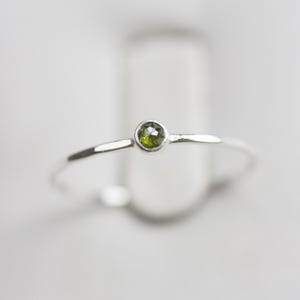 Peut inclure: Une bague en argent délicate ornée d'une petite pierre précieuse ronde vert olive. La pierre est sertie dans un sertissage simple et la monture est fine et polie. La bague est présentée sur un fond flou.