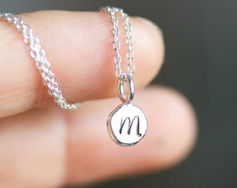 May include: A silver chain necklace with a small, round pendant featuring the letter "M" engraved on it.