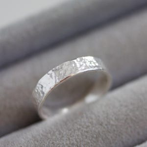 May include: A close-up of a silver ring with a hammered texture. The ring has a band with a slightly uneven surface, reflecting light. The ring is set against a soft grey background, highlighting its metallic sheen. A piece of jewellery.