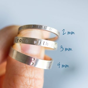 Peut inclure: Trois bagues en argent empilées les unes sur les autres. La bague du haut est gravée du mot "FREE". La bague du milieu est gravée des mots "strong with this". La bague du bas est gravée de deux petits points. Les bagues sont étiquetées avec leur largeur en millimètres : 2 mm, 3 mm et 4 mm.