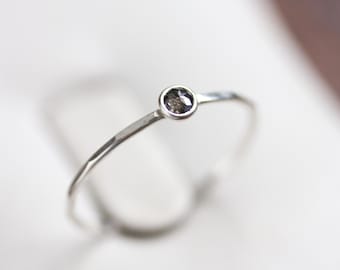 Peut inclure: Une fine bague en argent ornée d'une petite pierre précieuse ronde et gris foncé. La bague a un anneau fin et un design minimaliste. La pierre précieuse est sertie dans un sertissage à lunette, ajoutant une touche d'élégance.