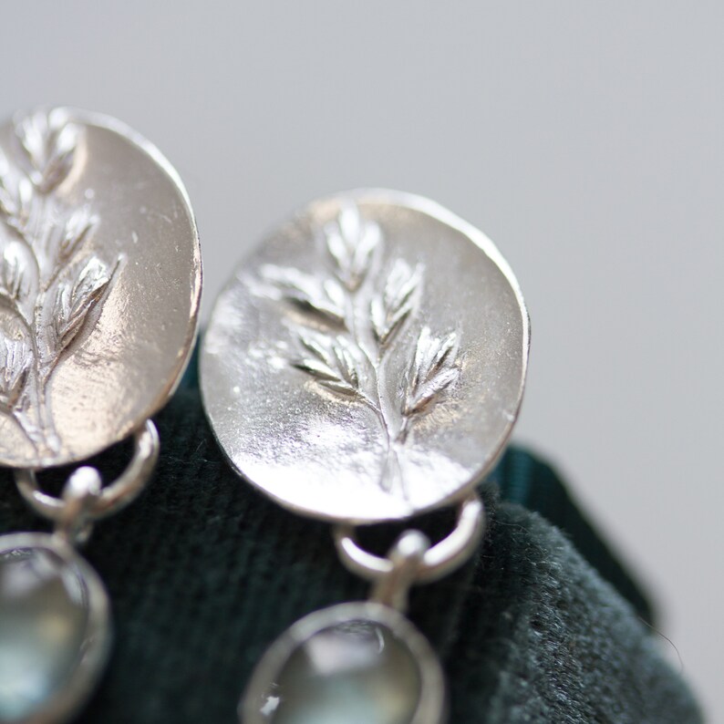 May include: A pair of silver earrings with a delicate branch design and a light blue gemstone dangle. The earrings are on a dark green velvet surface.