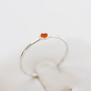 Könnte beinhalten: Ein zarter Silberring mit einem kleinen, runden, orangefarbenen Edelstein. Der Ring hat ein dünnes Band mit strukturiertem Design. Der Edelstein ist in einer einfachen Lünettenfassung gefasst, was einen minimalistischen und eleganten Look erzeugt.