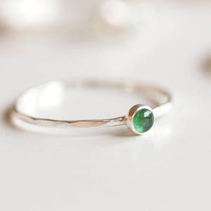 Könnte beinhalten: Ein zarter Silberring mit einem kleinen, runden, leuchtend grünen Edelstein. Der Ring hat ein schmales, strukturiertes Band. Der Edelstein ist in einer einfachen Fassung gefasst, die seine Farbe und Form hervorhebt. Minimalistisches und elegantes Design.