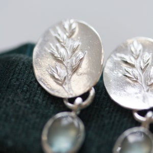 May include: Silver earrings with a delicate leaf design and a clear stone dangle. The earrings are on a dark green velvet background.