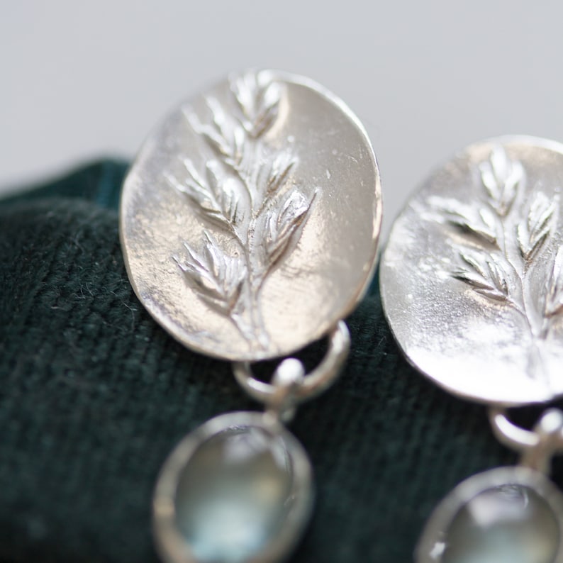 May include: Silver earrings with a delicate leaf design and a clear stone dangle. The earrings are on a dark green velvet background.