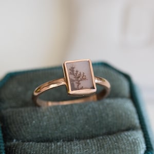 May include: A rose gold ring with a rectangular, light-coloured stone featuring a tree-like pattern. The ring is set in a simple, elegant design, showcasing the unique natural detail of the stone. The ring is displayed on a dark green velvet surface.