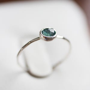 May include: A delicate silver ring featuring a faceted, oval-shaped teal gemstone. The gemstone is set in a bezel setting, and the ring has a thin, hammered band. The ring is shown against a soft, white background.