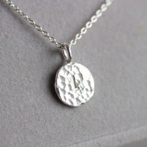 Pode incluir: Um colar com pingente de prata com um berloque circular martelado com a letra "L". O pingente pende de uma corrente fina. O colar é exibido em um fundo cinza.