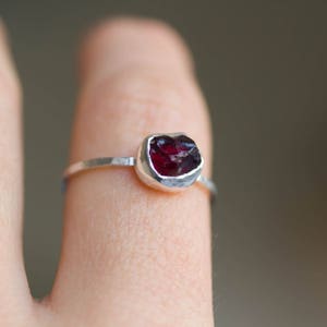 Könnte beinhalten: Ein silberner Ring mit einem roten Edelstein, der in einer Zargenfassung gefasst ist. Der Ring befindet sich an einem Finger.