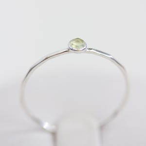 Op de afbeelding: Een zilveren ring met een kleine, lichtgroene edelsteen. De ring heeft een dunne, getextureerde band. De edelsteen is rond en gezet in een minimalistische stijl. Het ontwerp is eenvoudig en elegant, geschikt voor dagelijks gebruik.