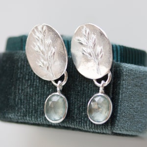 May include: A pair of silver earrings with a delicate leaf design and a light blue gemstone dangle. The earrings are on a dark green velvet background.