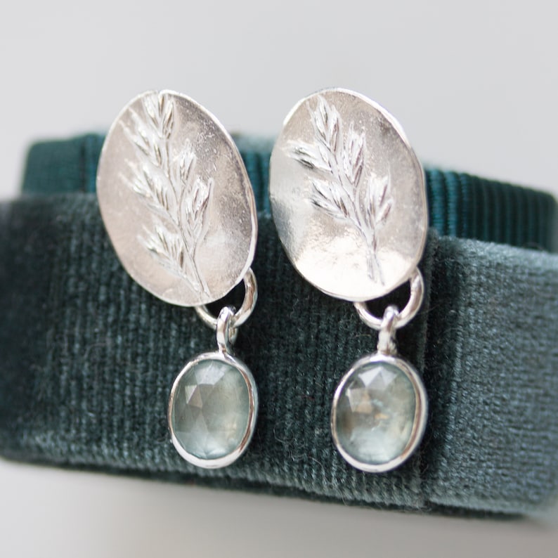 May include: A pair of silver earrings with a delicate leaf design and a light blue gemstone dangle. The earrings are on a dark green velvet background.