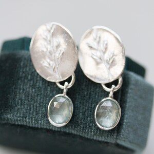 May include: A pair of silver earrings with a delicate branch design and a light blue gemstone dangle. The earrings are displayed on a dark green velvet surface.