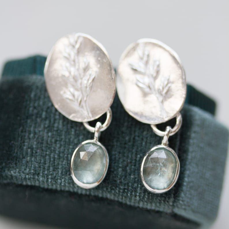 May include: A pair of silver earrings with a delicate branch design and a light blue gemstone dangle. The earrings are displayed on a dark green velvet surface.