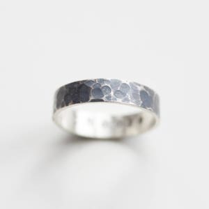 Op de afbeelding: Een zilveren ring met een gehammerde textuur. De band is breed en heeft een donkergrijze afwerking.